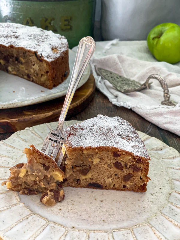 There is a slice of apple cake in the foreground with a cake fork standing up beside it and a piece of cake removed. In the background there is the rest of the cake off to one side, a vintage cake server on top of a napkin and a green apple in the background.